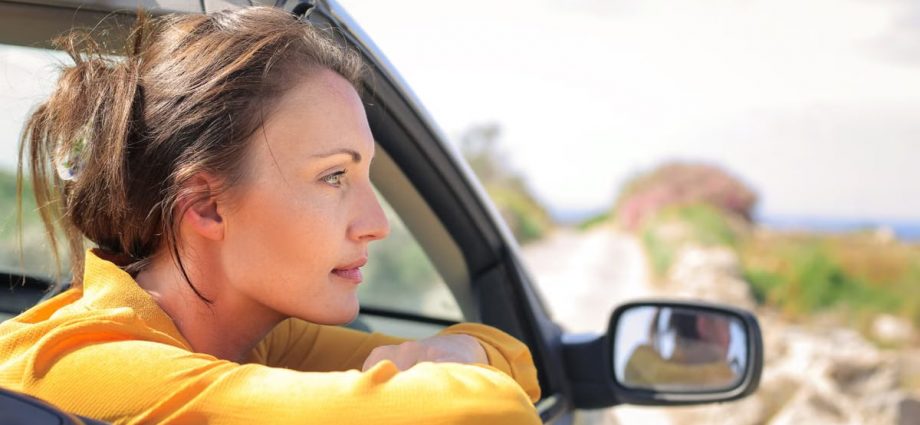 Profile view of a woman leaning out a convertible car enjoying a sunny day on a scenic road.